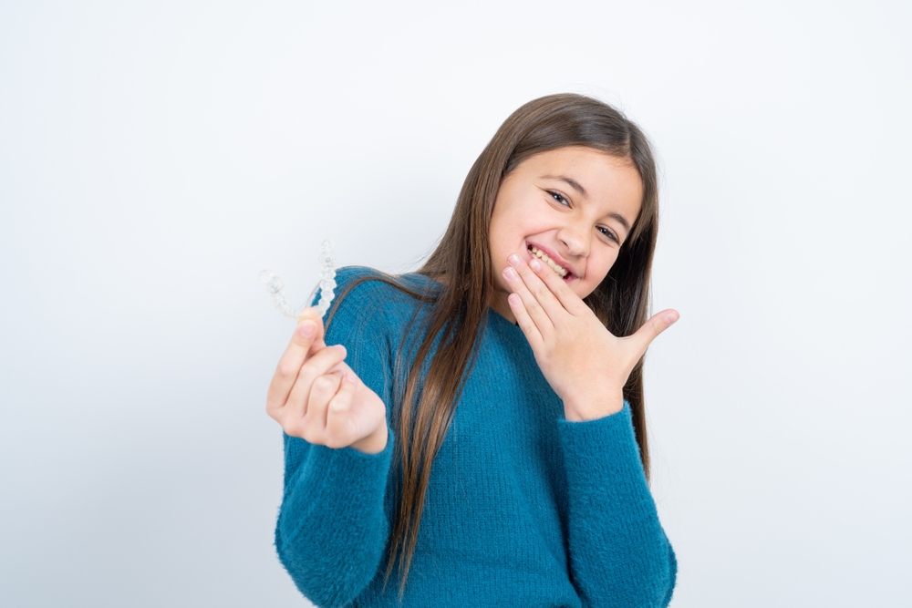Young Girl Holding Clear Aligners and Smiling A happy young girl holding clear aligners while showing a confident smile, representing orthodontic care for kids and teens