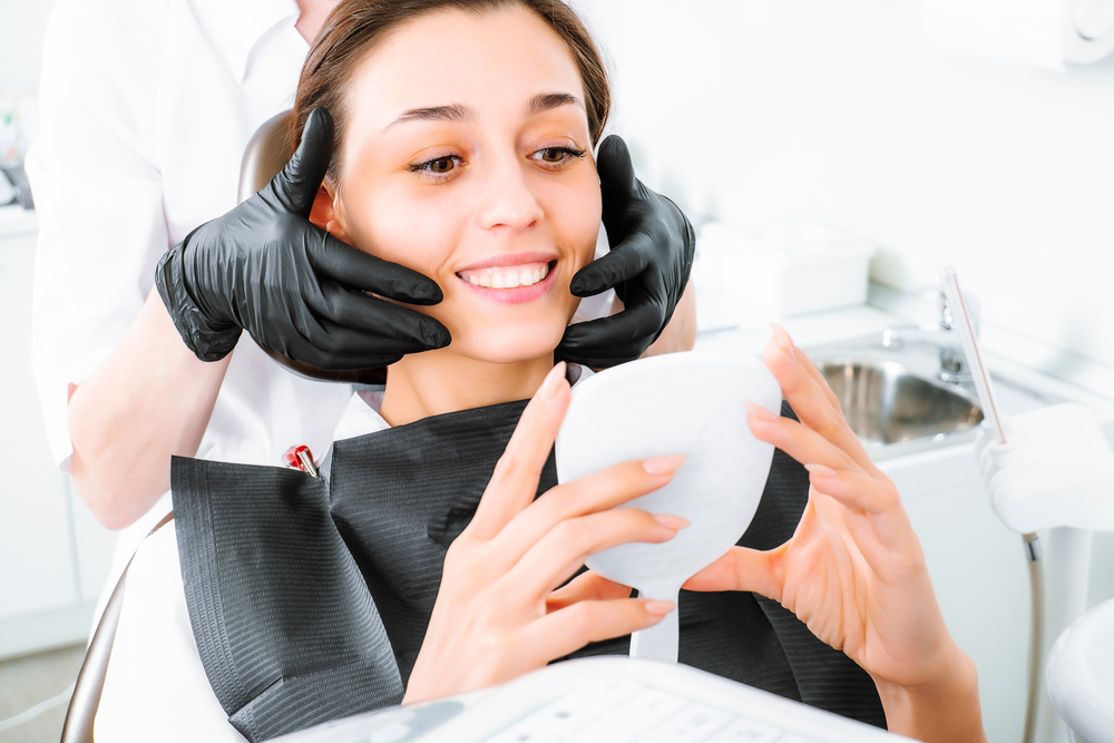 Patient Reviewing Veneer Results in Mirror - Porcelain Veneers Orange County A happy patient smiles while checking her enhanced teeth after cosmetic veneer treatment in a modern dental office - Porcelain Veneers Orange County