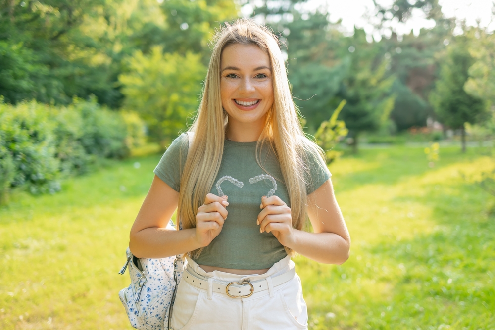 Clear Aligners for Straighter Smiles – Invisalign Irvine A young woman holding Invisalign-style aligners while smiling outdoors in natural light – Invisalign Irvine