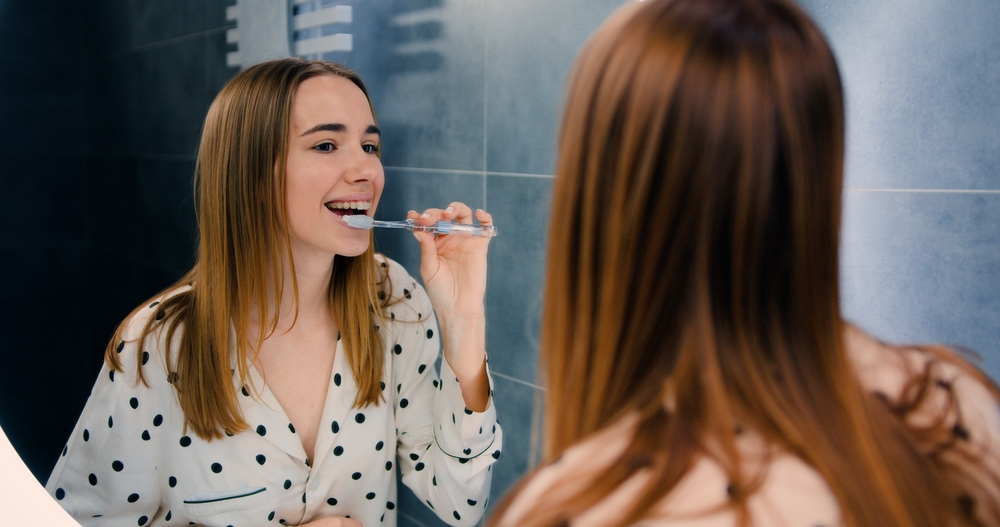 Daily Smile Care and Cosmetic Confidence - Porcelain Veneers Orange County A young woman brushes her teeth in the mirror, supporting healthy habits that complement porcelain veneer results - Porcelain Veneers Orange County